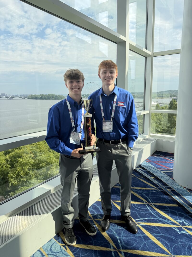 Two BT Students in blue dress shirts and gray pants stand indoors by large windows, smiling and holding a trophy together. Both wear name badges and event lanyards, celebrating their TSA Championships win as sunlight streams in over trees and water outside.