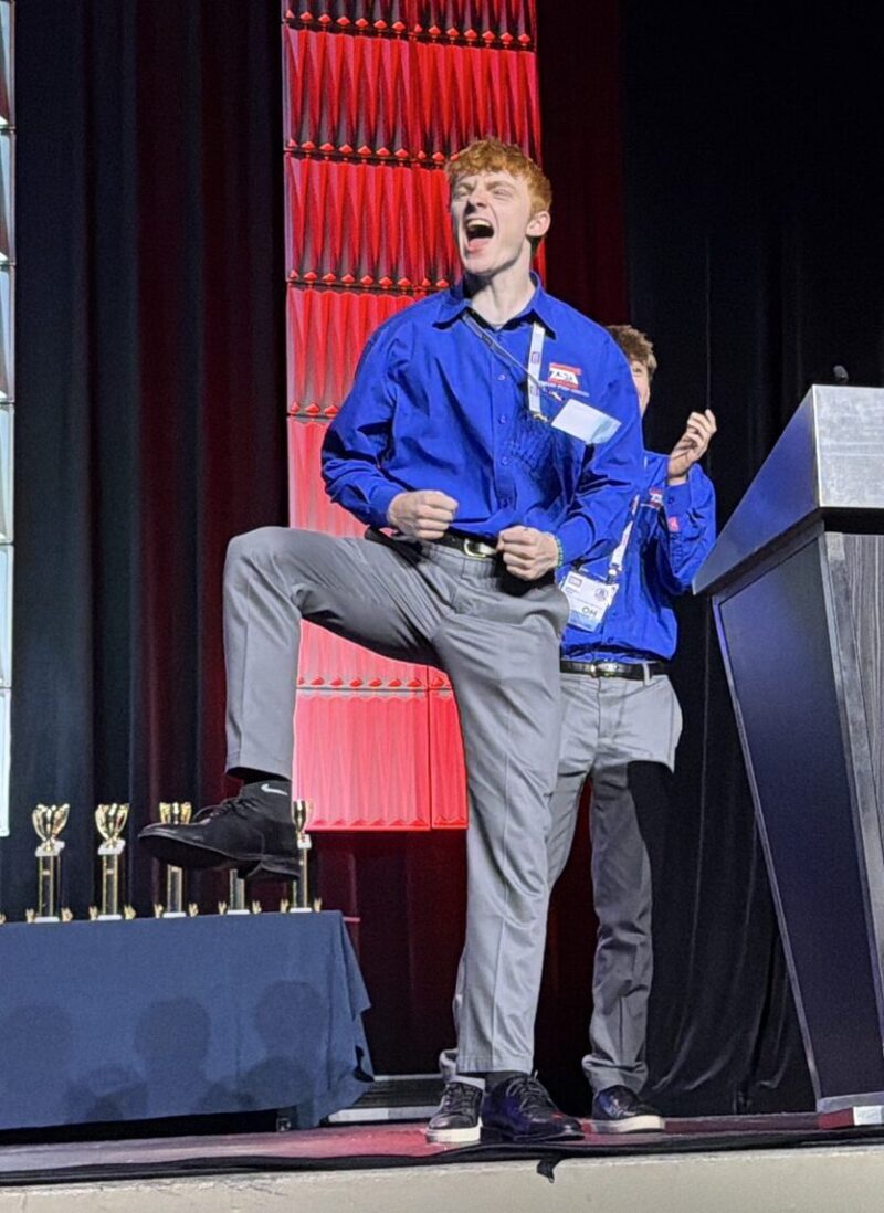 A young man in a blue dress shirt excitedly poses with one leg raised on stage next to a podium with a TSA Championships logo. Trophies are displayed in the background as other BT Students in matching shirts stand nearby at this national event.