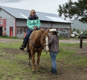 Lynsey Witt, featured in our Alumni Spotlight, wears sunglasses and a teal jacket as she sits on a brown horse while another woman in a cowboy hat stands beside them, holding the reins. They are outside near a barn on a grassy area.