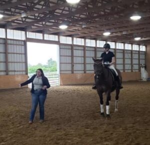 Alumni Spotlight: Lynsey Witt stands in an indoor riding arena beside a helmeted rider on a dark horse, reflecting her passion for Equine Science. The arena features dirt flooring, wooden beams, and large windows that fill the space with natural light.
