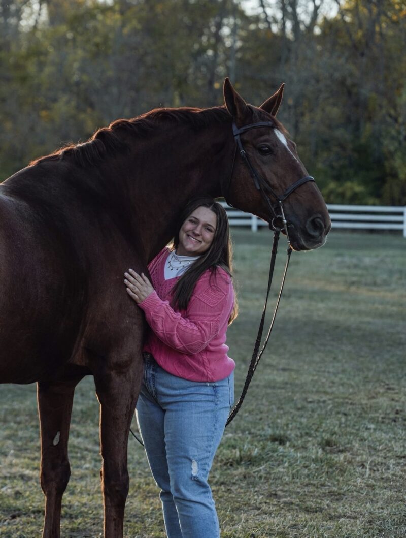 A woman in a pink sweater and blue jeans stands outdoors, smiling and hugging a large brown horse. Highlighting her passion for Equine Science, this Alumni Spotlight captures Lynsey Witt against grass, trees, and a white fence in soft evening light.
