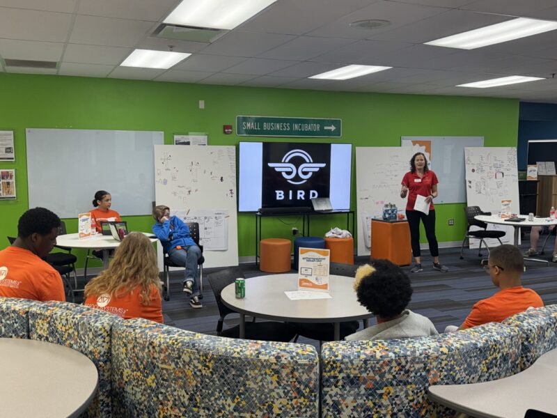 A woman stands and speaks to a group of young people in orange shirts seated around a classroom. A large screen displays the Bird logo, and there are whiteboards and colorful decor in the background.
