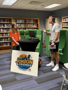 Two people speak into headsets at a table with laptops in a library. A large sign in front of them reads "Jon Jon & Friends Cincinnati" with a microphone image. Bookshelves fill the background.