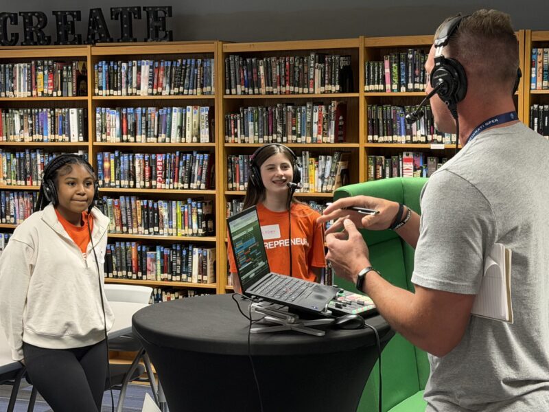 A man wearing headphones talks to two girls in orange shirts standing in front of a laptop and microphone in a library, with bookshelves and a 