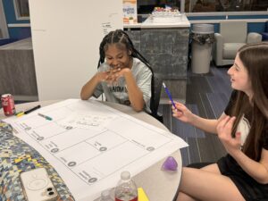 Two girls sit at a round table working on a large worksheet. One holds a pen and gestures while the other looks thoughtful. Various items, including a drink, phone, and markers, are on the table in a casual, modern room.