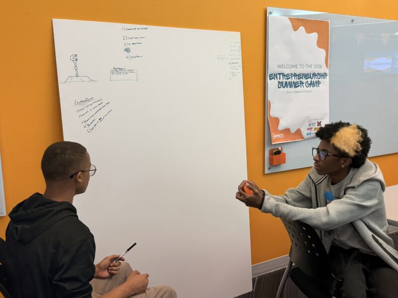 Two students sit facing each other in a classroom, discussing and taking notes on a large whiteboard. A poster on the wall reads 