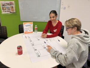 Two young people sit at a round table, working on a business model canvas worksheet. One wears a red shirt, the other a gray hoodie. There are drinks, paper, and pens on the table. A green wall and whiteboard are in the background.