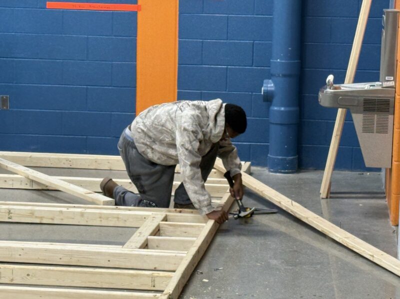 A construction student in a gray hoodie kneels on the floor, working with a tool on a wooden frame structure inside a room with blue and orange walls.