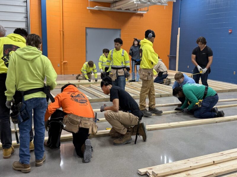 A group of Construction Students in work clothes measure and arrange wooden beams on a shiny indoor floor, likely participating in a construction or carpentry workshop. The walls are painted orange and blue.