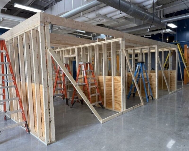 Partially constructed wooden framing for a room or building, supported by braces, with several colored ladders around it. The scene is indoors with exposed pipes and ducts on the ceiling—a hands-on space for construction students to learn.
