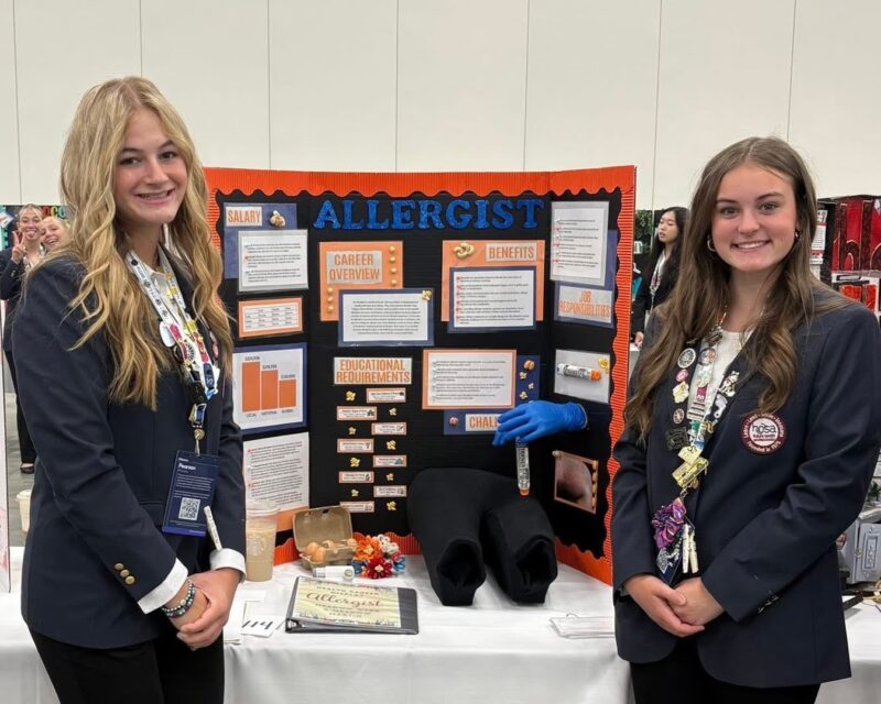 Two young women stand smiling in front of a trifold display board titled 
