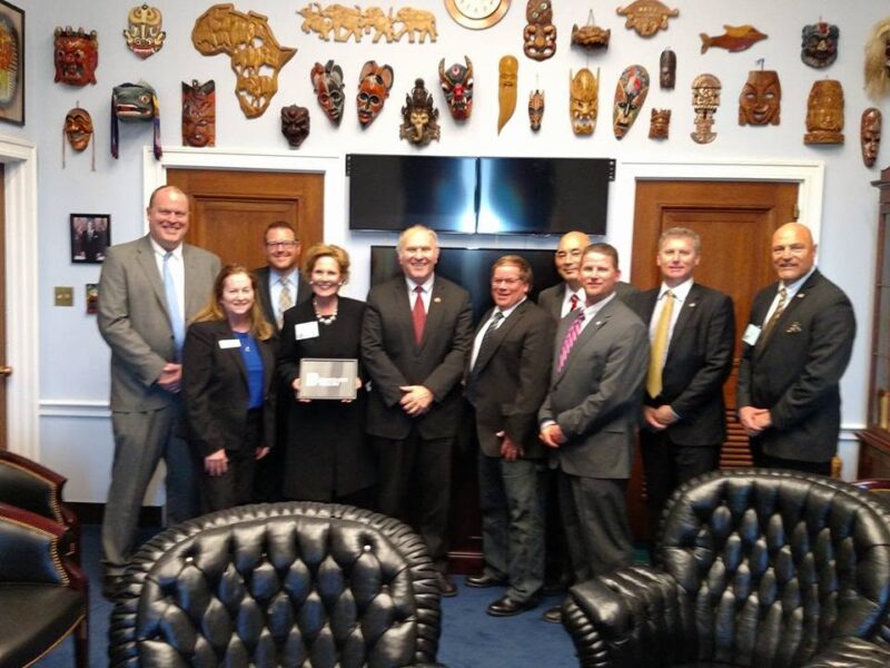 A group of eleven people in business attire, including Jim Miller, pose for a photo in an office with many decorative masks on the walls, leather chairs, and a TV behind them at Butler Tech.