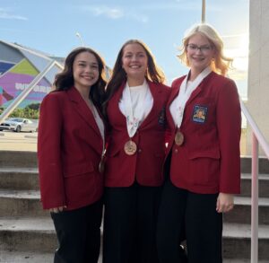 Three young women, SkillsUSA students wearing red blazers and medals, stand smiling on outdoor steps. A large stadium with a colorful mural and “FIFA World Cup 2026” text is visible as the sun sets behind them, hinting at their national recognition.