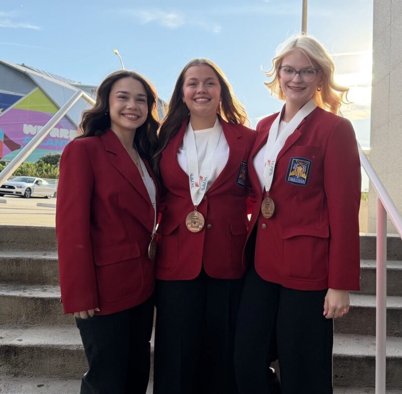 Three young women, SkillsUSA students wearing red blazers and medals, stand smiling on outdoor steps. A large stadium with a colorful mural and “FIFA World Cup 2026” text is visible as the sun sets behind them, hinting at their national recognition.