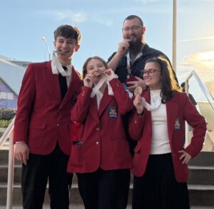 Four students stand on outdoor stairs, smiling and posing with medals. Three wear matching red blazers, proudly representing SkillsUSA, while the fourth stands behind in a dark shirt. A colorful building is in the background.