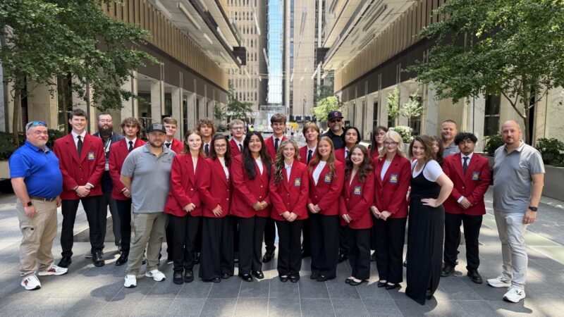 SkillsUSA students in red blazers and black pants stand in rows outside between tall buildings, with several adults in casual attire on either side. Trees line the walkway as the group smiles at the camera, celebrating national recognition.