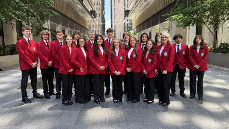 A group of 17 people stands outdoors in two rows, all wearing matching red blazers, black pants, and white shirts. Smiling at the camera, they proudly represent SkillsUSA student achievement in an urban setting with trees and glass buildings.
