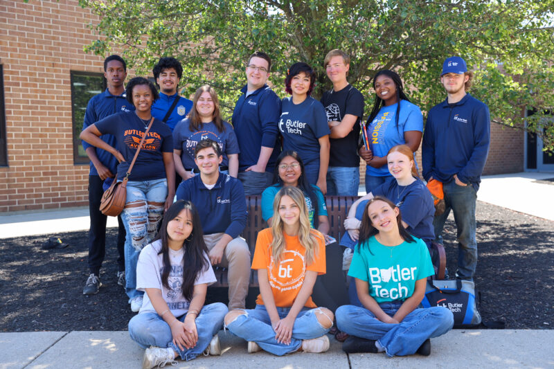 A group of sixteen diverse Student Ambassadors in casual Butler Tech shirts and jackets pose and smile outdoors in front of a brick building and a leafy tree. Some are seated on a bench, while others stand behind them.