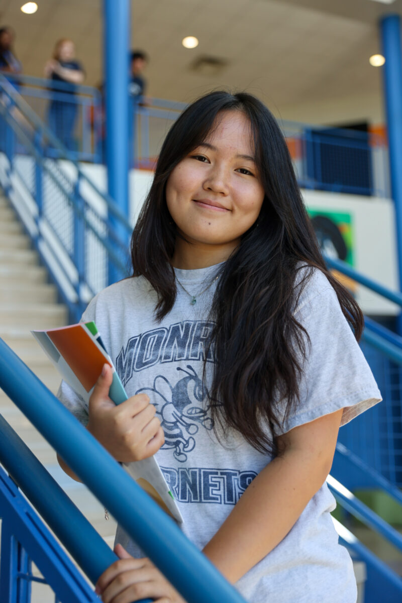 A young woman with long dark hair, one of the campus representatives, holds notebooks and smiles while standing by a blue railing in a school hallway, with stairs and other students in the background.