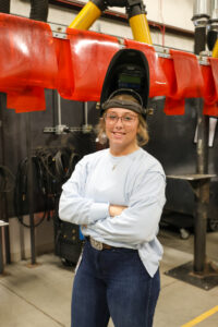 Alexia Morgan, a college student, stands with arms crossed in an industrial workshop, smiling at the camera. Wearing glasses and a welding helmet flipped up, she is dressed in a light blue shirt and jeans with welding equipment behind her.
