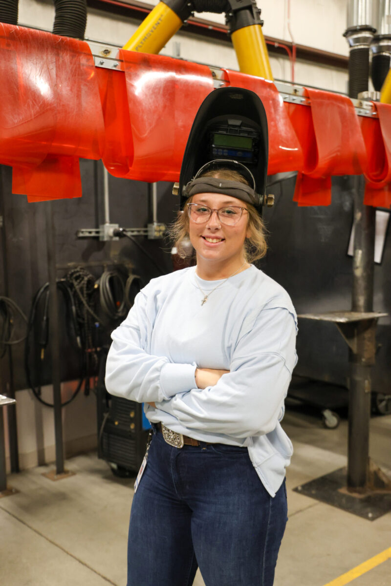 Alexia Morgan, a college student, stands with arms crossed in an industrial workshop, smiling at the camera. Wearing glasses and a welding helmet flipped up, she is dressed in a light blue shirt and jeans with welding equipment behind her.