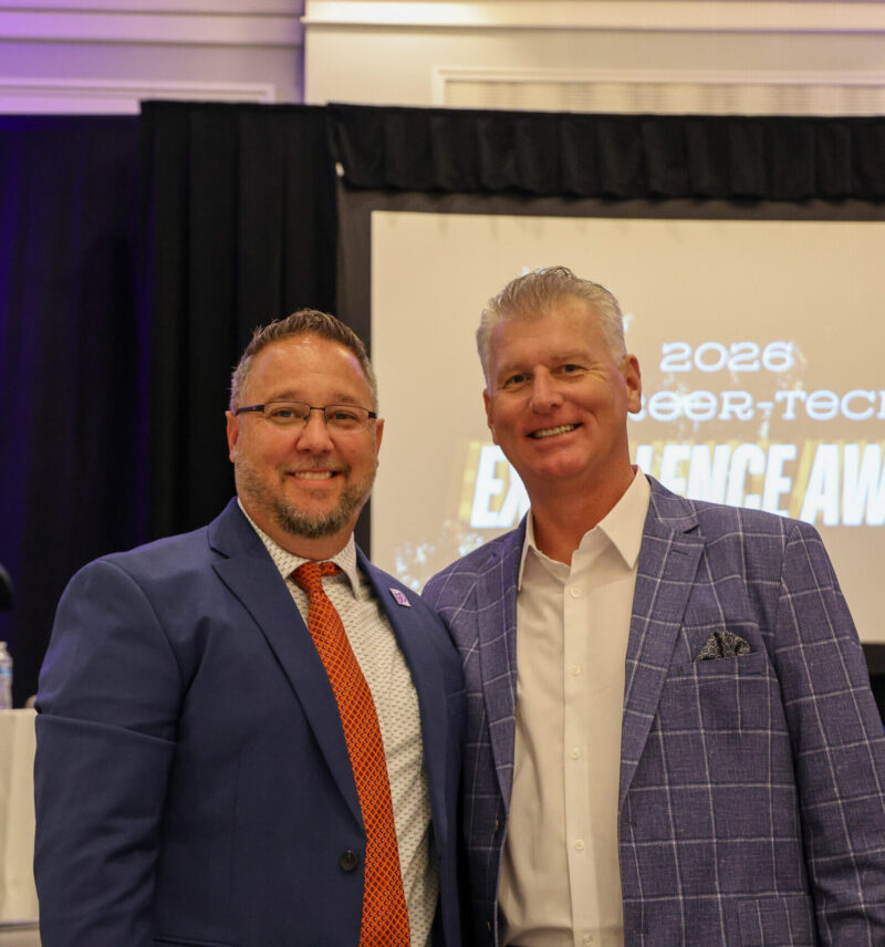 Two men in suits smile at an indoor event celebrating education honors. Behind them, a screen displays 
