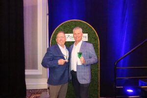 Two men in blue blazers stand and smile in front of a green backdrop, one holding a green drink and the other pointing towards him. A sign behind them highlights Ohio ACTE education honors, with hints of blue lighting and a mention of Butler Tech setting the scene in the background.