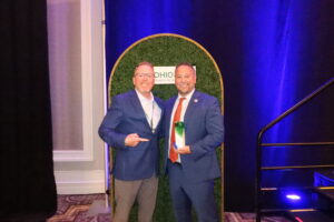 Two men in suits smile for a photo; one holds an education honors award and the other points at him. They stand in front of a green backdrop with a sign that reads "OHIO" and the logo for Ohio ACTE, celebrating achievements at Butler Tech. Blue lighting sets the scene.