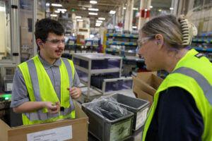 Two people in yellow safety vests and safety glasses talk at a workstation in a factory, surrounded by equipment and containers, with shelves and industrial machinery in the background—a scene highlighting the collaborative network fostered by Project LIFE.