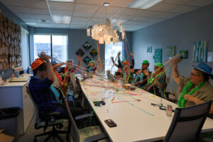 People wearing colorful hats and beads sit around a conference table, tossing colorful streamers in the air as they celebrate the successes of Project LIFE in a decorated office meeting room filled with bright window light, reflecting the vibrant energy of their network.