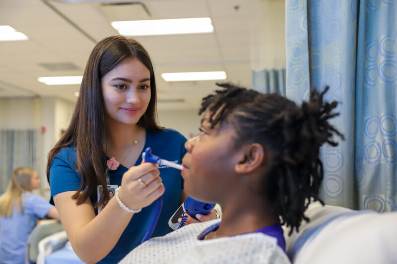 A nurse in blue scrubs smiles while checking a patient’s temperature with an oral thermometer as the patient sits in a hospital bed, demonstrating the compassionate care typical of Licensed Practical Nursing (LPN) professionals.