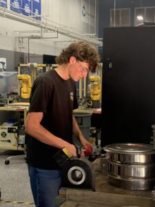 A young man wearing safety glasses uses a power tool on a metal part in a workshop, with robotic arms and equipment visible in the background—a dynamic scene that showcases the spirit of mechatronics within a student-led enterprise.