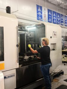 A person wearing gloves operates a Milltronics CNC machine in a mechatronics workshop, adjusting a large metal piece. Banners hang on the wall in the background, reflecting the innovative spirit of the student-led enterprise.
