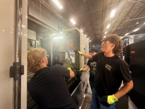 Three people wearing safety glasses and gloves work together, inspecting machinery in a well-lit industrial setting. As part of a student-led enterprise, one person points a flashlight at the equipment while the others observe closely, showcasing their collaborative approach to mechatronics.