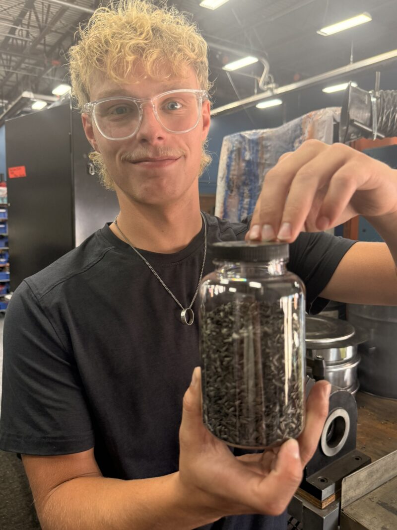 A person wearing clear safety glasses and a black shirt holds up a jar filled with small black metal shavings in an industrial workshop setting, highlighting the hands-on experience gained through a student-led enterprise focused on mechatronics.
