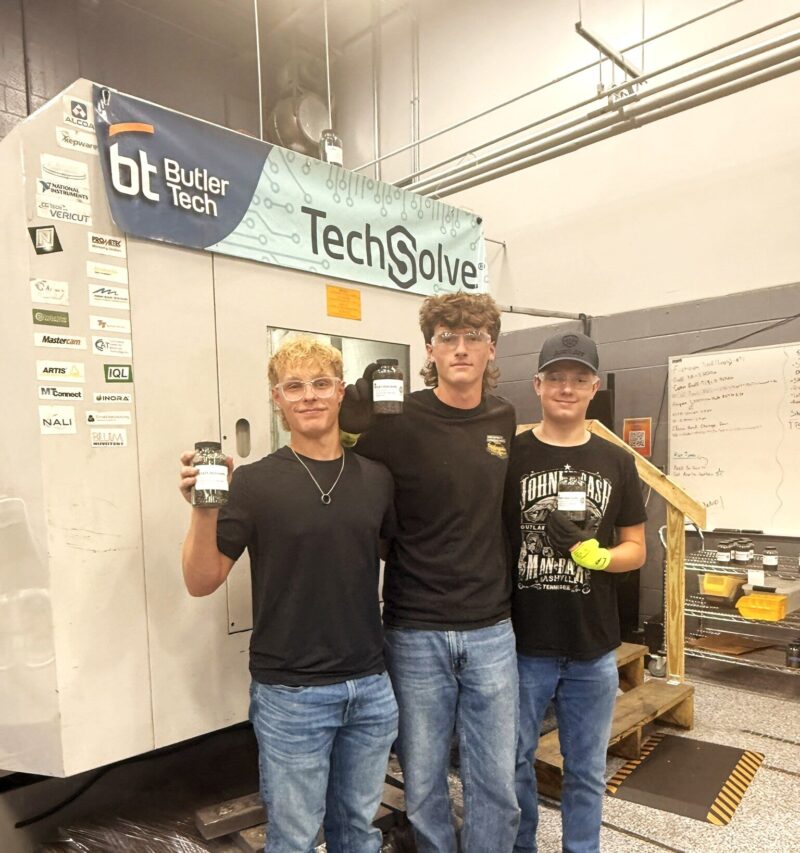 Three young men stand in a workshop, smiling and holding objects. Behind them is a large machine labeled “TechSolve” and “Butler Tech” with various stickers, reflecting their involvement in student-led enterprise and mechatronics. Shelving and equipment are visible in the background.