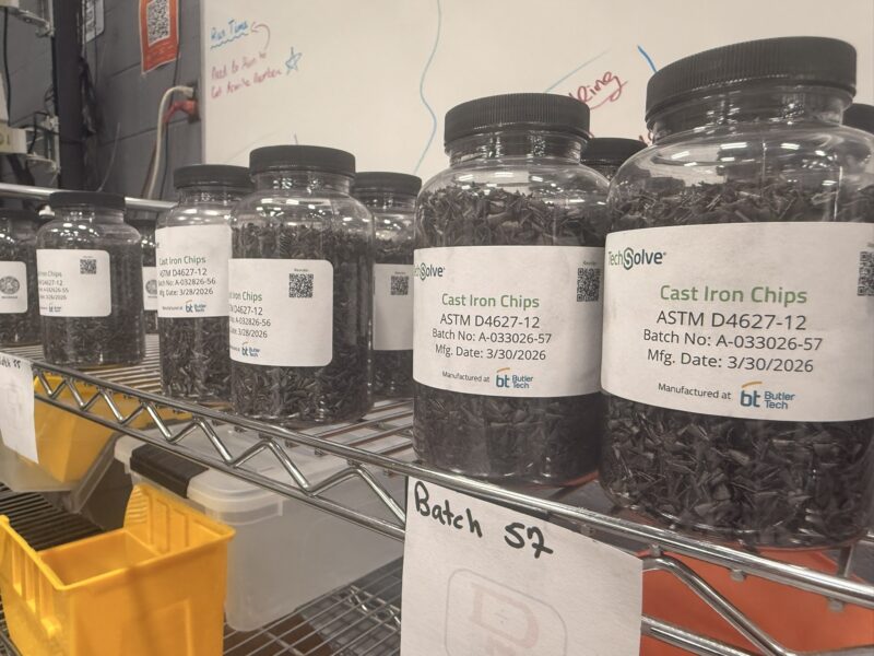 Several jars labeled “Cast Iron Chips” sit on a metal shelf, each with batch and manufacturing dates. A “Batch 57” sign is visible below the shelf in a lab or workshop setting, reflecting the meticulous work of a student-led enterprise focused on mechatronics.