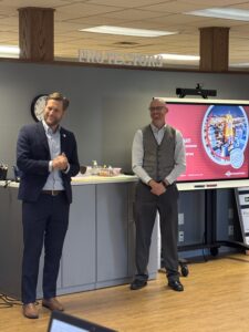Two men stand and talk in an office setting near a screen displaying a colorful presentation. One man gestures while speaking, and the other stands with hands clasped, smiling. A “PROTECTORS” sign is visible above them, while information about regional opportunities in partnership with Butler Tech and REDI Cincinnati appears on the screen.