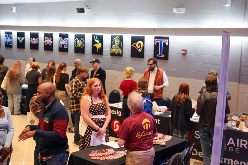 People stand and talk at a career fair with various school banners hanging on the wall behind them. Tables have informational brochures and candy. The room is bright and filled with SOA students of different ages.
