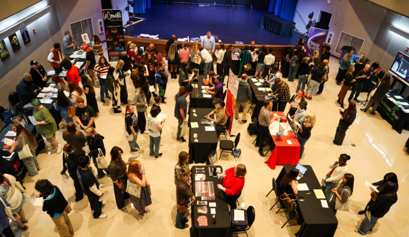 A large group of SOA students stand and walk around tables at a busy career fair, interacting with vendors and each other in a spacious hall while showcasing their professional skills.