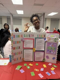 A smiling Middletown student stands behind a science fair board titled “Donning Safety Stack,” with instructions for protective gear and sample items arranged on a table for career technical education.