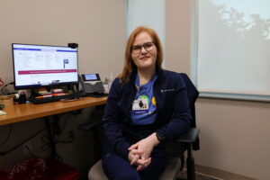 A woman wearing glasses and a navy blue uniform sits in an office chair in front of a computer monitor.