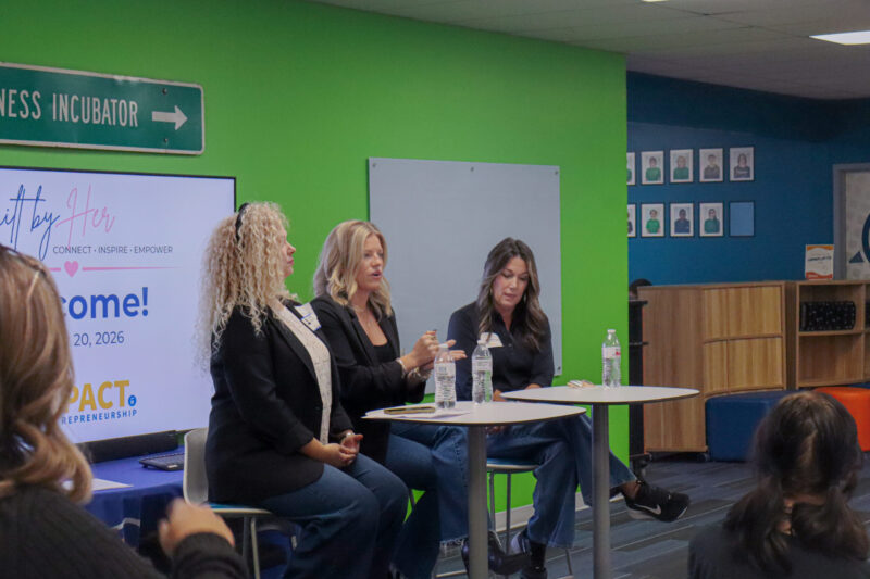 Three women sit at a leadership development panel discussion in front of a green wall, with a monitor to their left displaying “Gift by Her” branding.