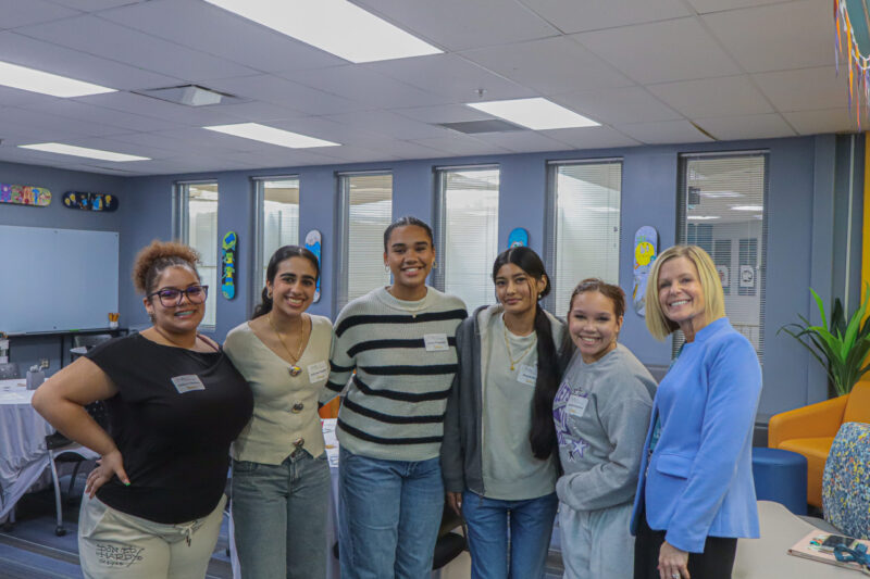 Five young women and an older woman smiling together in a classroom, embodying student inspiration and leadership.