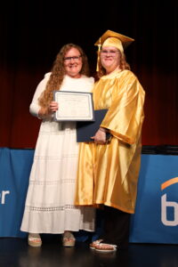 A woman in a white dress stands next to a graduate in a gold cap and gown, who is holding a medical assisting certificate.