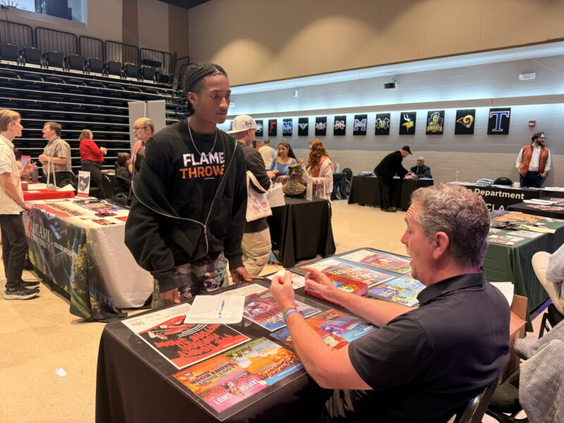 A SOA student talks to a man seated at a colorful table display during a school career fair in a gym, with other students and tables in the background, highlighting opportunities to develop professional skills.