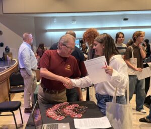 A man and a woman shake hands at a table with papers and flyers, while SOA students stand in line nearby at a career fair in a large, well-lit room, showcasing their professional skills.