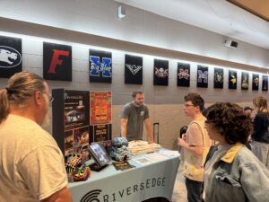 SOA Students stand at the RiversEdge booth, showcasing skull art and colorful flyers. School team banners hang above in the bright, casual Career Fair setting.