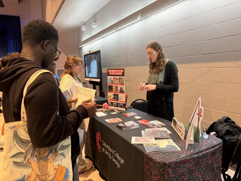 A group of SOA students talk to a representative at a table for Columbus College of Art & Design, looking at brochures and art samples during a career fair.