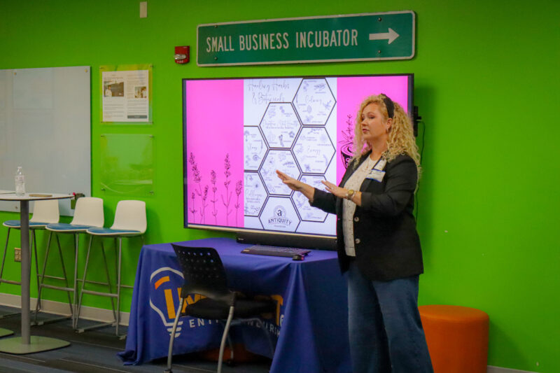 A woman, embodying student inspiration, stands before a digital screen displaying a “Healing Herbs & Botanicals” infographic in a room with a green wall and a “Small Business Incubator” sign.